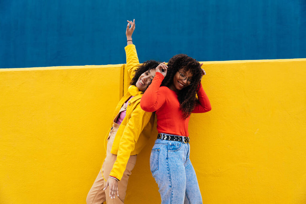 Young happy women dancing and having fun outdoor. Teenagers listening to music with smartphone and headphones in a yellow and blue modern urban area - Foto, afbeelding