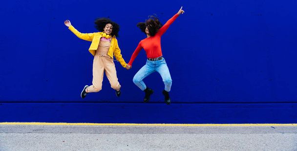 Young happy women dancing and having fun outdoor. Teenagers listening to music with smartphone and headphones in a yellow and blue modern urban area - Fotografie, Obrázek