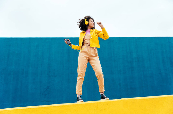 Young happy woman dancing and having fun outdoor. Teenager listening to music with smartphone and headphones in a yellow and blue modern urban area - Foto, immagini