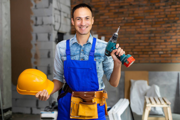 Porträt eines positiven, gut aussehenden jungen männlichen Bauarbeiters mit Hut, der in die Kamera lächelt und Trockenbau hält, während er auf der Baustelle arbeitet - Foto, Bild
