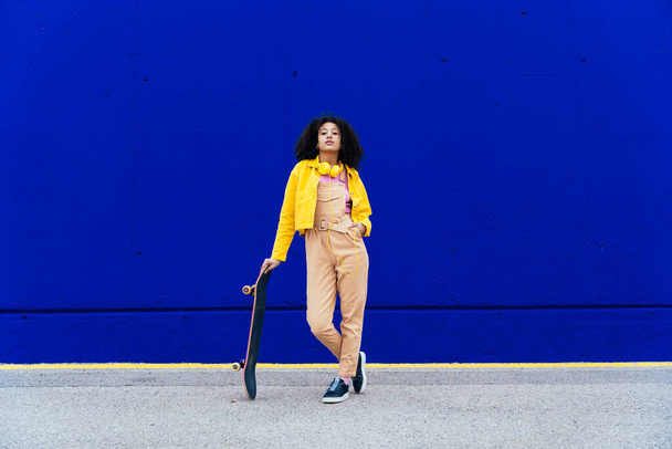 Young happy woman having fun outdoor with her skateboard. Teenager listening to music with smartphone and headphones in a yellow and blue modern urban area - Fotó, kép