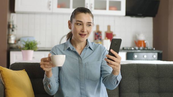 Жінка переглядає соціальні мережі, дивиться відео, дивиться фотографії в соціальних мережах, п'є каву, сидить на дивані у вітальні з мобільним телефоном в руці
. - Фото, зображення