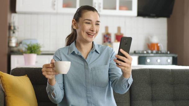 Жінка, яка серфінгує в соціальних мережах, переглядає відео, дивиться на фотографії в соціальних мережах, п'є каву, сидить на дивані у вітальні зі своїм мобільним телефоном, дуже здивована новинами, які вона бачить
. - Фото, зображення
