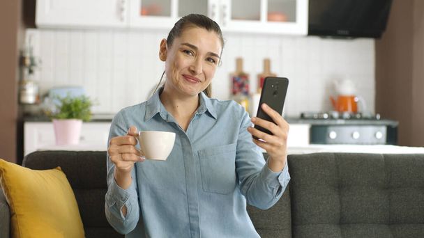 Femme naviguant sur les réseaux sociaux, regardant des vidéos, regardant des photos sur les réseaux sociaux, buvant du café, assise sur un canapé dans le salon avec un téléphone portable à la main, souriant et regardant la caméra. - Photo, image