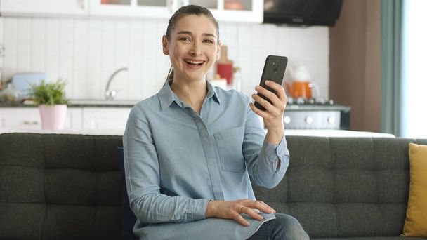 The young woman was stunned by the news she saw while surfing the internet on her phone. Surprised by news on smartphone, woman smiles at camera. - Photo, Image