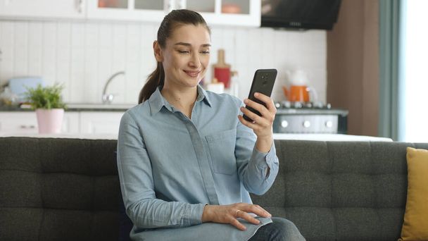 Young woman reading news or text on the phone at home. The young woman laughs at the message she sees. Woman browsing the internet, browsing social media accounts. - Photo, Image