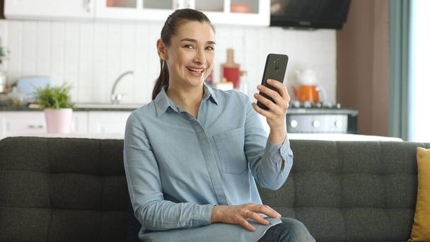 The young woman was stunned by the news she saw while surfing the internet on her phone. Surprised by news on smartphone, woman smiles at camera. - Foto, afbeelding