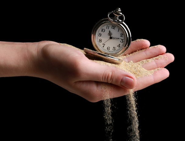 Ασημένιο ρολόι τσέπης στα χέρια και της άμμου που ρέει μακριά σε μαύρο φόντο - Φωτογραφία, εικόνα