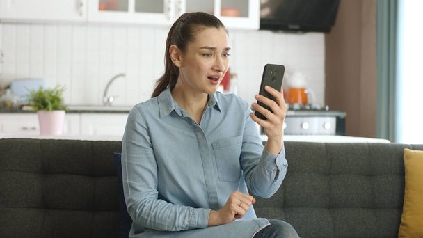 Houkutteleva nuori nainen käyttää älypuhelinta istuessaan sohvalla. Nuori nainen on hyvin järkyttynyt ja vihainen uutisista, joita hän näkee älypuhelimessaan. Surullisten uutisten käsite Internetissä. - Valokuva, kuva