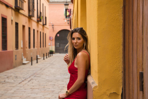 Young, blonde, beautiful woman in a red dress is visiting seville. The woman poses for the camera very elegant and like a model in the typical streets of the city. Holidays and travels - Foto, Imagem