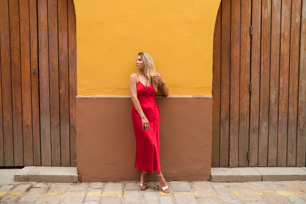 Young, blonde, beautiful woman in a red dress is visiting seville. The woman is posing for the camera very elegant for the photos. The woman has a yellow wall in the background. Holidays and travels. - Fotografie, Obrázek