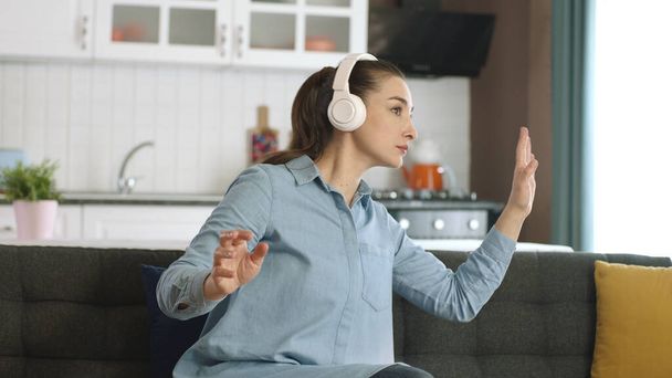 Young happy woman sitting on sofa at home, listening to music and singing with headphones. He greets his friend by waving his hand. Home entertainment concept. Portrait of happy fun woman. - Photo, Image