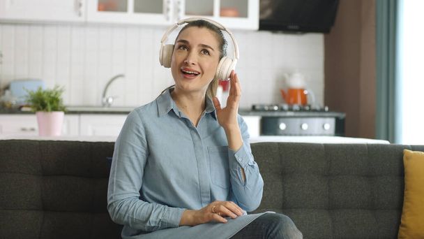 Молода щаслива жінка сидить на дивані вдома, слухає музику з навушниками та співає. Концепція домашніх розваг. Щасливий жіночий портрет
. - Фото, зображення