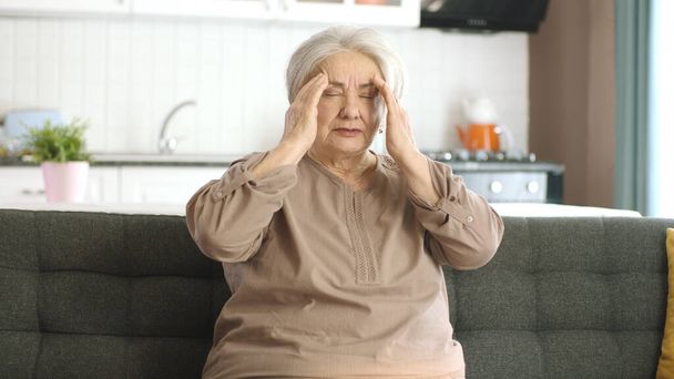 Staruszka odczuwa silny ból w głowie. Staruszka siedzi na krześle i boli ją głowa. Kobieta masująca swoją bolącą głowę rękami. Portret starszej kobiety z problemami zdrowotnymi. - Zdjęcie, obraz