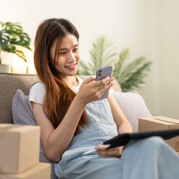 Jonge ondernemer controleert online bestellingen op smartphone terwijl hij comfortabel op de bank zit en met tablet werkt in de woonkamer op kantoor. - Foto, afbeelding