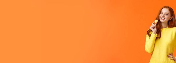 Талия портрет прекрасная рыжая девушка держит вкусный желтый макарон у рта и радостно улыбается, как едят сладости, купить десерт для романтического свидания валентинки, позируя веселый оранжевый фон
. - Фото, изображение