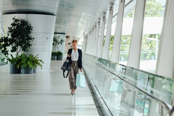 Mujer de negocios feliz sosteniendo bolsas de compras y usando un teléfono móvil mientras camina en una pasarela - Foto, Imagen