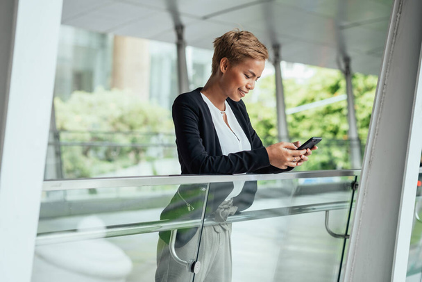 Χαμογελαστή επιχειρηματίας πληκτρολογώντας μήνυμα κειμένου στο smartphone της, ενώ ακουμπά σε γυάλινο κιγκλίδωμα σε μια σύγχρονη βεράντα - Φωτογραφία, εικόνα