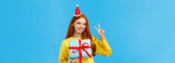 Pas-up portrét roztomilé šťastné zrzky žena obdrží dárek v krabici, slaví vánoční svátky, na sobě maškarní Nový rok klobouk a ukazuje mír znamení, radostně se usmívá, modré pozadí. - Fotografie, Obrázek