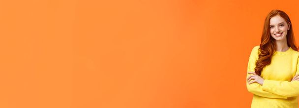 Vacances, personnes et concept d'hiver. Jolie jeune fille souriante aux cheveux roux en pull jaune debout dans une posture puissante et confiante, caméra souriante, bras croisés sur la poitrine, fond orange
. - Photo, image