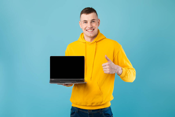 Happy caucasian man showing laptop computer empty screen and gesturing thumbs up, standing on blue background. Freelancer guy advertising great website or distance job offer - Foto, immagini