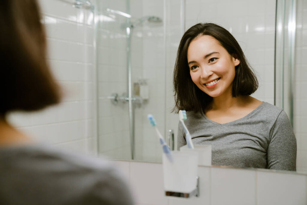 Young smiling asian girl standing in front of a mirror in the bathroom at home - Fotoğraf, Görsel