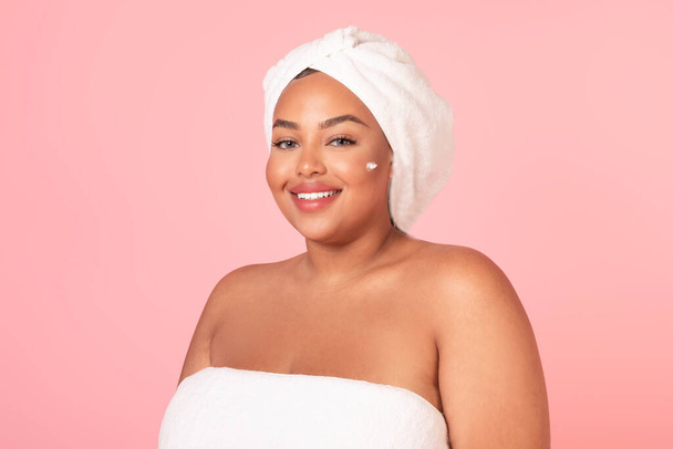 Beauty day concept. Portrait of beautiful black obese woman with cream on cheek and white towel on head smiling at camera, applying hydration moisturizer on face, pink studio background - Foto, imagen