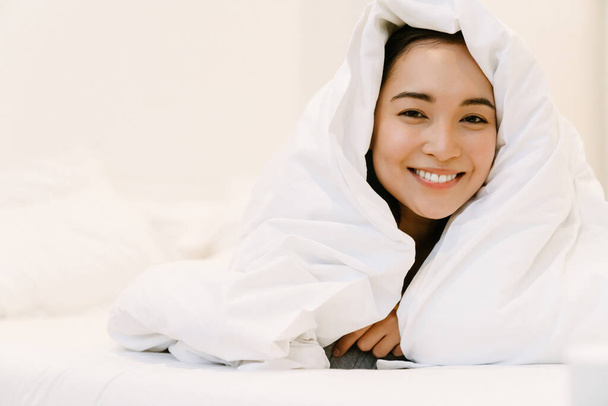 Young smiling asian girl in home suit lying on bed under covers at home - Foto, Imagem