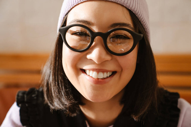 Young asian woman in eyeglasses smiling and looking at camera indoors - 写真・画像