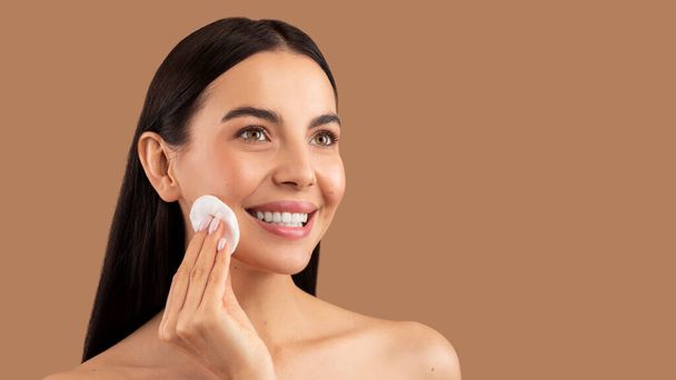 Topless long-haired brunette pretty young lady cleansing face skin with micellar water and cotton pad, looking at copy space for advertisement and smiling, beige studio background, panorama - 写真・画像