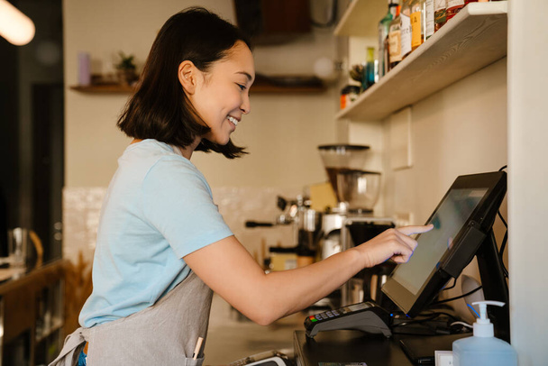 Asian barista woman wearing apron smiling while working with cash register in cafe - Zdjęcie, obraz