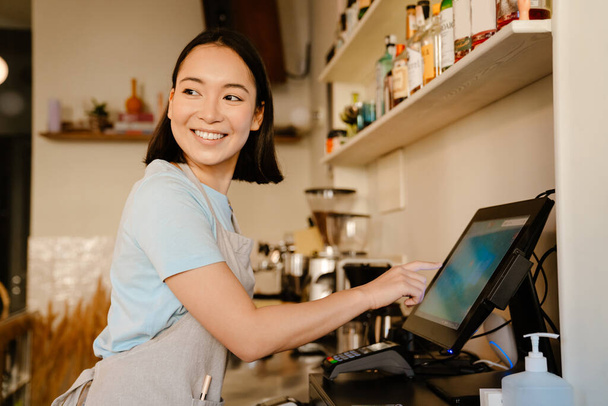 Asian barista woman wearing apron smiling while working with cash register in cafe - Фото, изображение