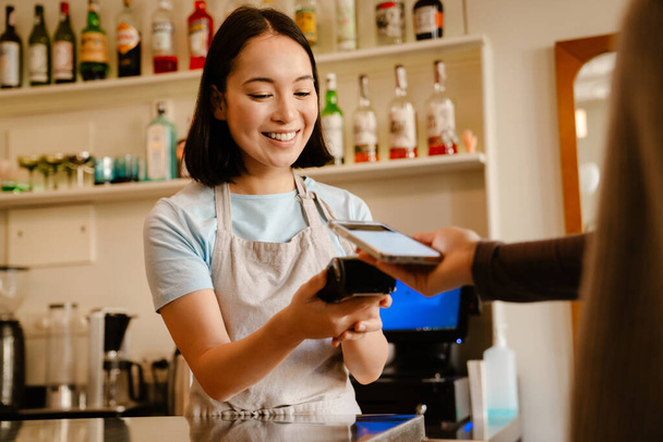 Asian barista woman wearing apron smiling while working with client in cafe - Φωτογραφία, εικόνα
