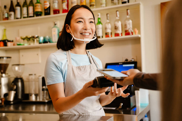 Asian barista woman in face mask smiling while working with client in cafe - Fotó, kép