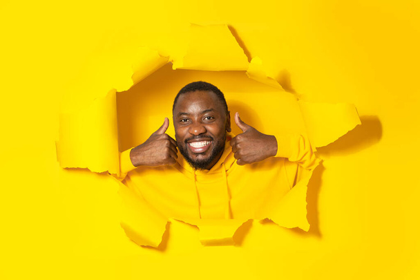 Vreugdevolle zwarte man gebaren duimen omhoog en glimlachen naar de camera door gat in gescheurd geel papier, goedkeuring van groot aanbod, poseren in de studio. Ik hou van het concept - Foto, afbeelding