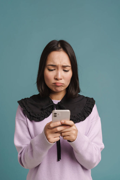 Young asian woman frowning and using mobile phone isolated over blue background - Foto, afbeelding
