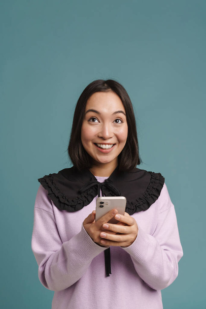 Młoda azjatycka kobieta uśmiechnięta i korzystająca z telefonu komórkowego odizolowana na niebieskim tle - Zdjęcie, obraz