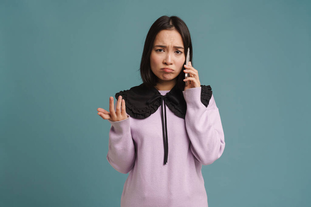 Young asian woman frowning and talking on mobile phone isolated over blue background - Fotografie, Obrázek
