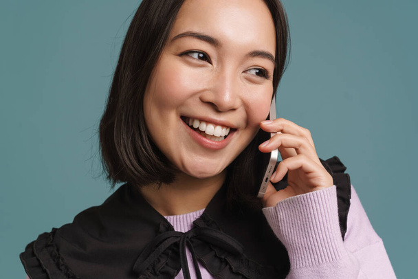 Young asian woman smiling and talking on mobile phone isolated over blue background - Photo, Image