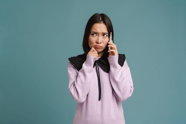 Young asian woman frowning and talking on mobile phone isolated over blue background - Fotó, kép