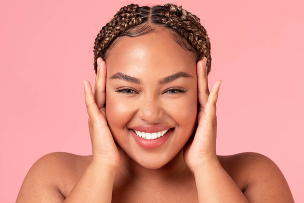 Retrato de la hermosa mujer afroamericana feliz tocando su cara y sonriendo a la cámara, posando sobre fondo de estudio rosa, primer plano - Foto, imagen