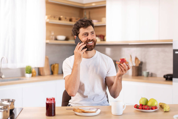 Blij volwassen blanke man met baard in wit t-shirt eet sandwich, telefoontjes per telefoon in moderne keuken interieur. Goedemorgen en ontbijt, communicatie, nieuws en roddels, geweldige aanbieding en advertentie thuis - Foto, afbeelding