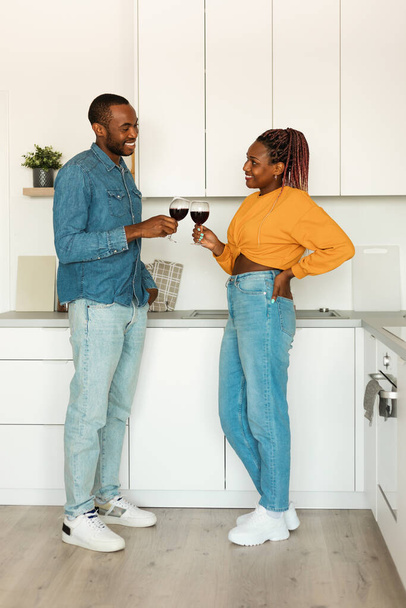Häusliche Romantik. Afroamerikanisches Paar mit Weingläsern, die im Kücheninneren stehen, liebende schwarze Männer und Frauen, die sich zu Hause verabreden, Zeit miteinander verbringen, Ganzkörperaufnahme - Foto, Bild