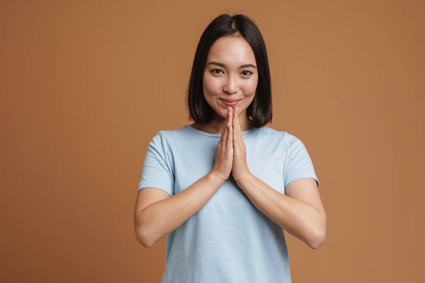 Young asian woman wearing t-shirt smiling while holding palms together isolated over beige background - Foto, imagen