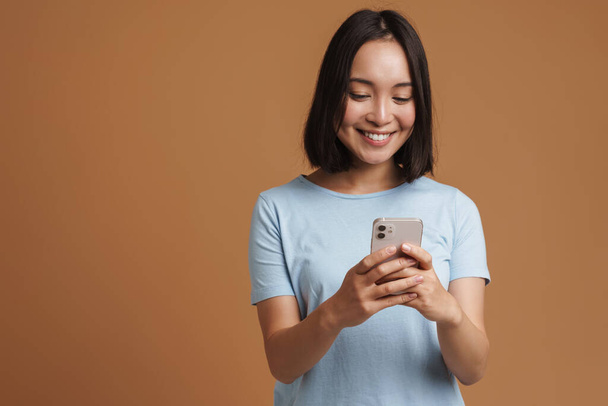 Young asian woman wearing t-shirt laughing while using mobile phone isolated over beige background - Fotó, kép