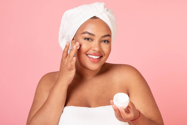 美しいアフリカ系アメリカ人の体の正の女性のクリームの瓶を保持し、ピンクの背景にポーズをとり、頬に保湿剤を適用します。毎日の治療、自宅での美容ケアのための化粧品 - 写真・画像