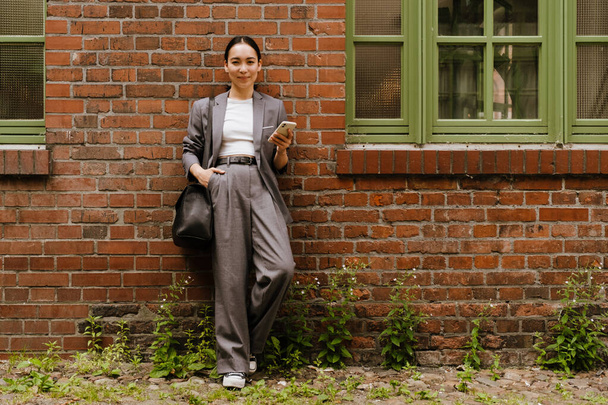 Asian young businesswoman using cellphone while standing by wall outdoors - Valokuva, kuva