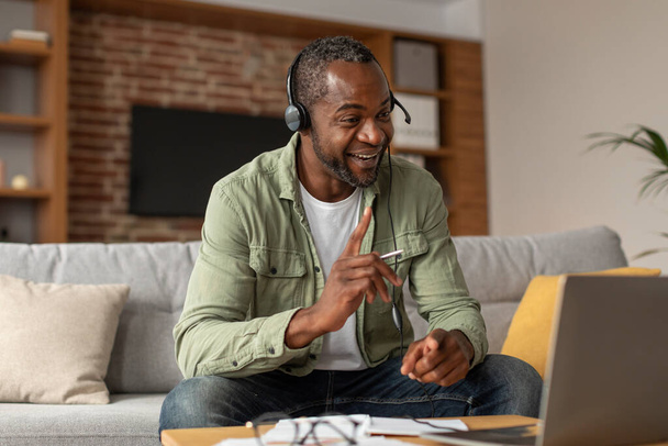 Boldog elfoglalt középkorú afro-amerikai férfi fülhallgatóval nézi a laptopot, gesztikulál, beszél az ügyféllel a nappali belsejében. Videó lecke, ügyfélszolgálat, munka és üzlet az otthoni irodában - Fotó, kép
