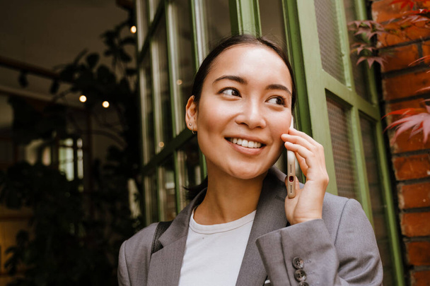 Asiática joven mujer de negocios sonriendo y hablando en el teléfono celular, mientras que de pie por la construcción de la calle - Foto, imagen