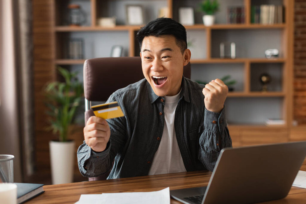 Удовлетворенный удивленный взволнованный взрослый японский бизнесмен с открытым ртом держит кредитную карту, сделать победный жест в интерьере домашнего офиса. Победа, успешная работа, удаленное финансирование бизнеса и эмоции - Фото, изображение